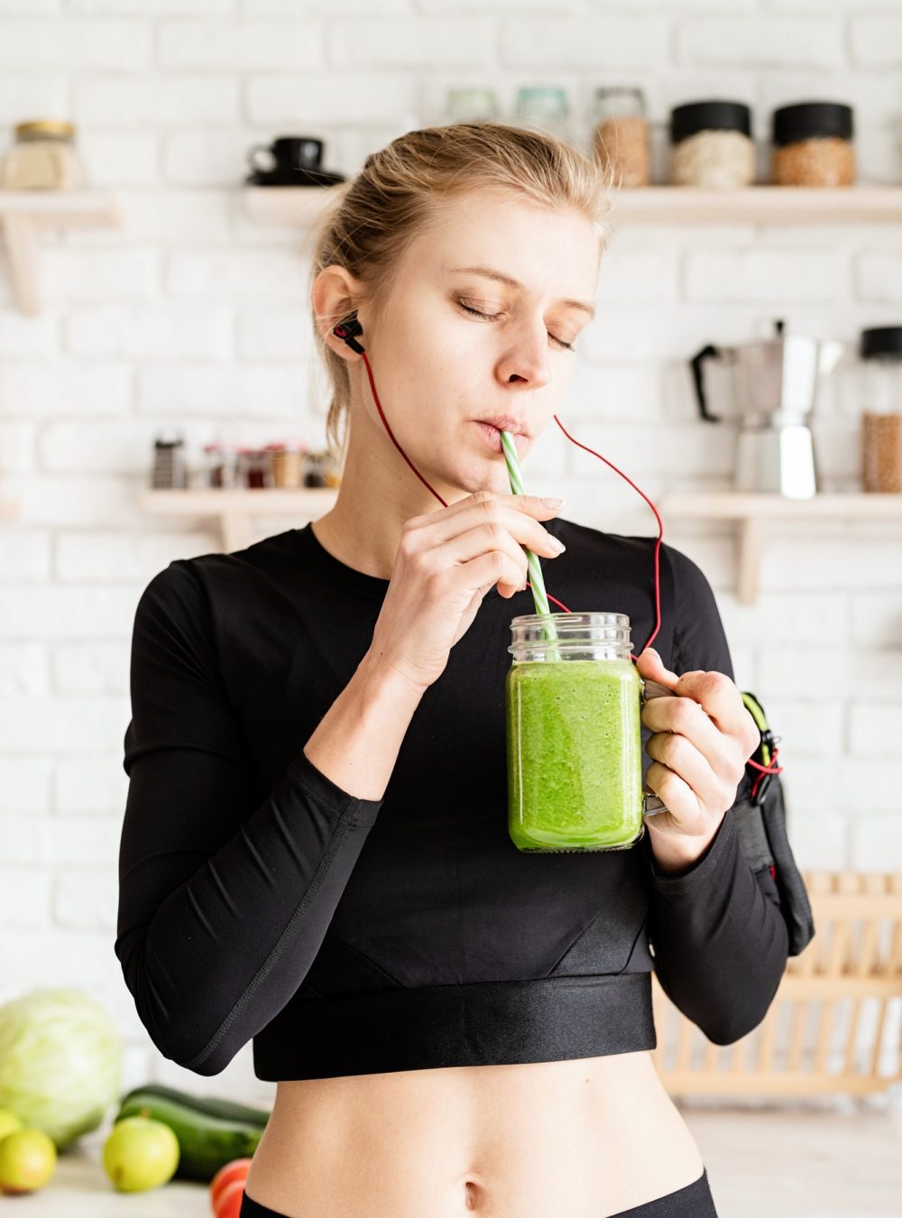 Mujer con ropa deportiva negra bebiendo un batido verde saludable en un frasco de vidrio con popote, en una cocina con fondo de pared de ladrillo blanco.