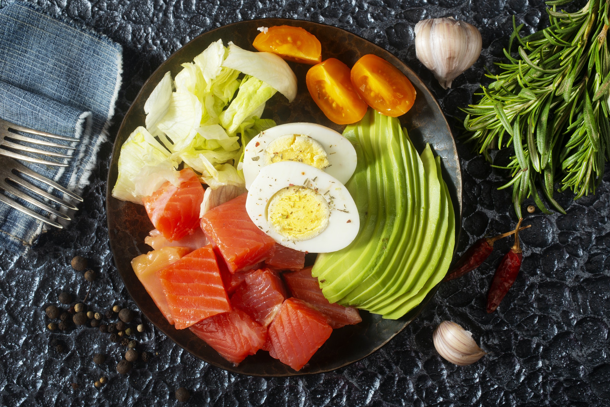 Salmón con huevo cocido - Beneficios de la nutrición funcional / Ensalada keto con salmón fresco en cubos, aguacate, huevo cocido, lechuga y tomates cherry, acompañada de ajo, romero y especias sobre fondo rústico.