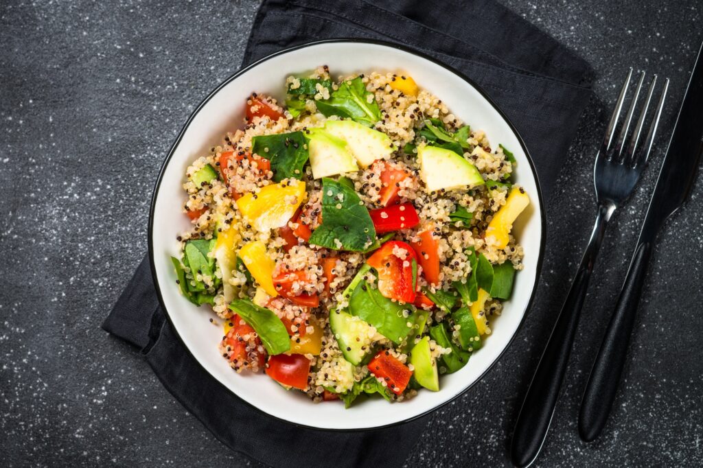 Ensalada de quinoa con espinaca, aguacate, pimientos de colores y tomate, servida en un bowl blanco sobre fondo oscuro.