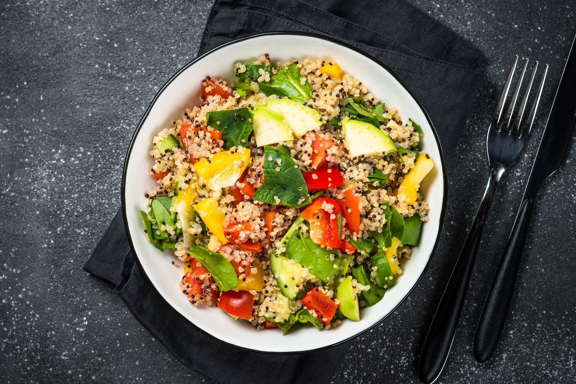 Ensalada de quinoa con espinaca, aguacate, pimientos de colores y tomate, servida en un bowl blanco sobre fondo oscuro.